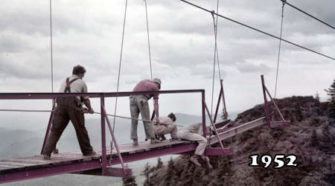 1952 Construction of the Original Mile High Swinging Bridge at Grandfather Mountain A historical photo from 1952 showing workers constructing the original Mile High Swinging Bridge at Grandfather Mountain, North Carolina. Three men are positioned on the partially completed bridge, working with cables and beams, with a backdrop of rugged mountain cliffs and cloudy skies.
