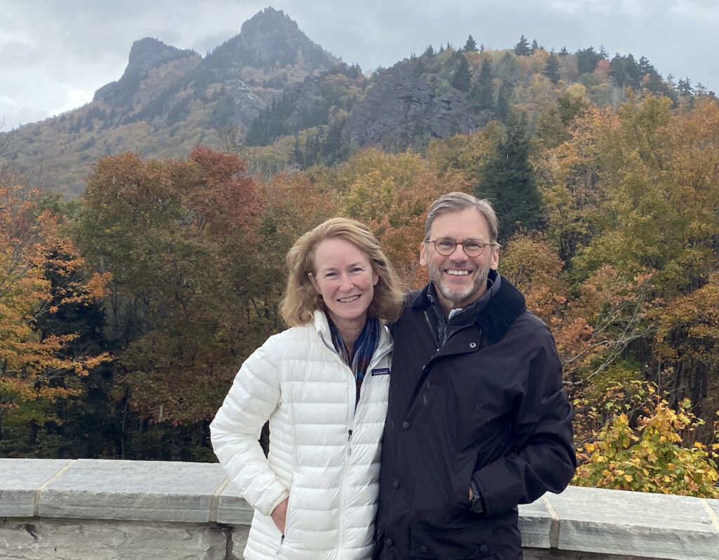 A woman and a man, Katherine and Tom Bunn, standing at Grandfather Mountain's scenic Half Moon Overlook, with Grandfather Mountain's peaks in the background.