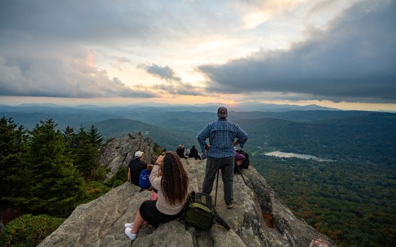 A group of people sit on a rocky ledge at Grandfather Mountain looking at a sunset over the mountains.